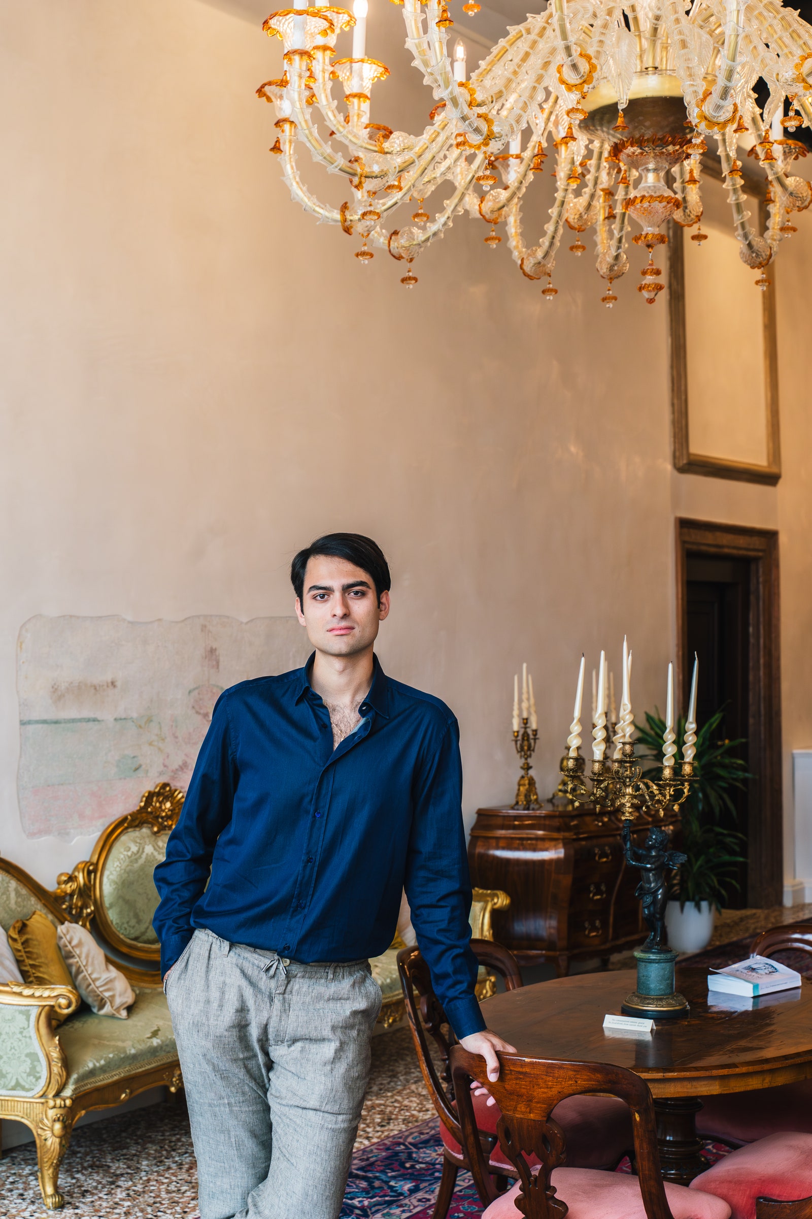 תמונה עשויה להכיל מנורה נברשתית אדם למבוגר מכנסי ביגוד ספרים ריהוט ושולחן ריהוט ושולחן