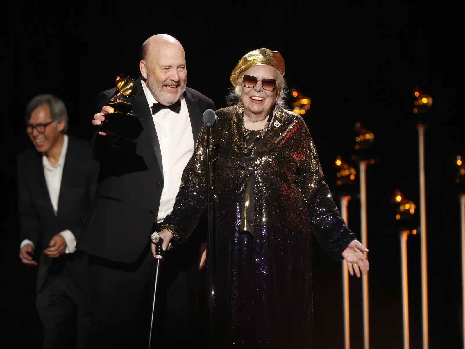 התמונה עשויה להכיל את ג'וני מיטשל ביגוד מעיל פנים ראש אדם צילום דיוקן תאורה לבוש וחליפה רשמיים